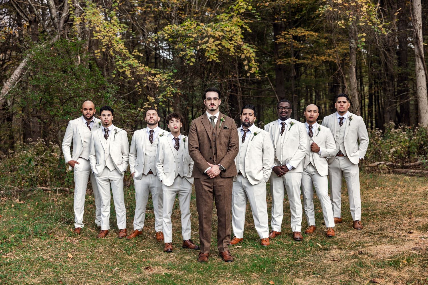 Groom in brown suit stands confidently with groomsmen in matching ivory suits in an outdoor fall setting.
