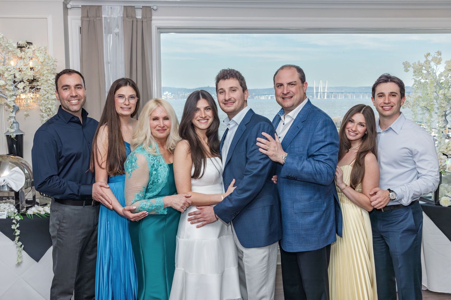 Family group of eight posing in formal attire with a scenic waterfront view behind.