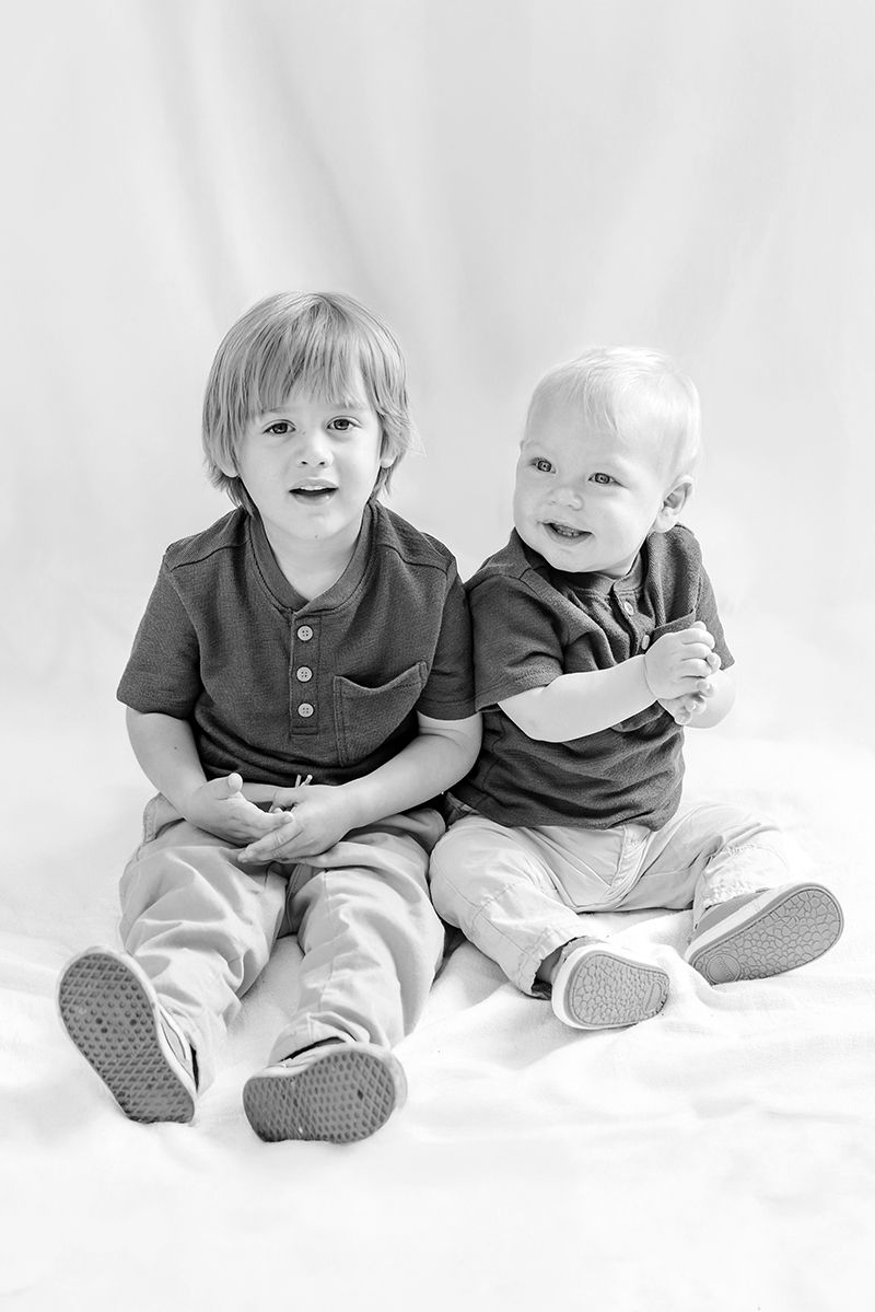 Black and white portrait of two young brothers sitting together smiling on white backdrop.