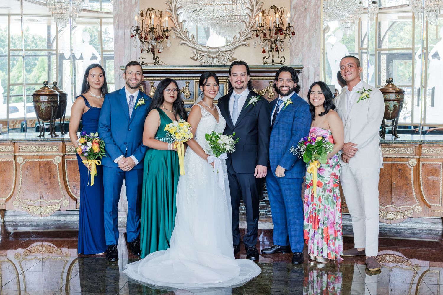 Wedding party group portrait with bride, groom, and attendants in blue and green gowns in elegant venue.