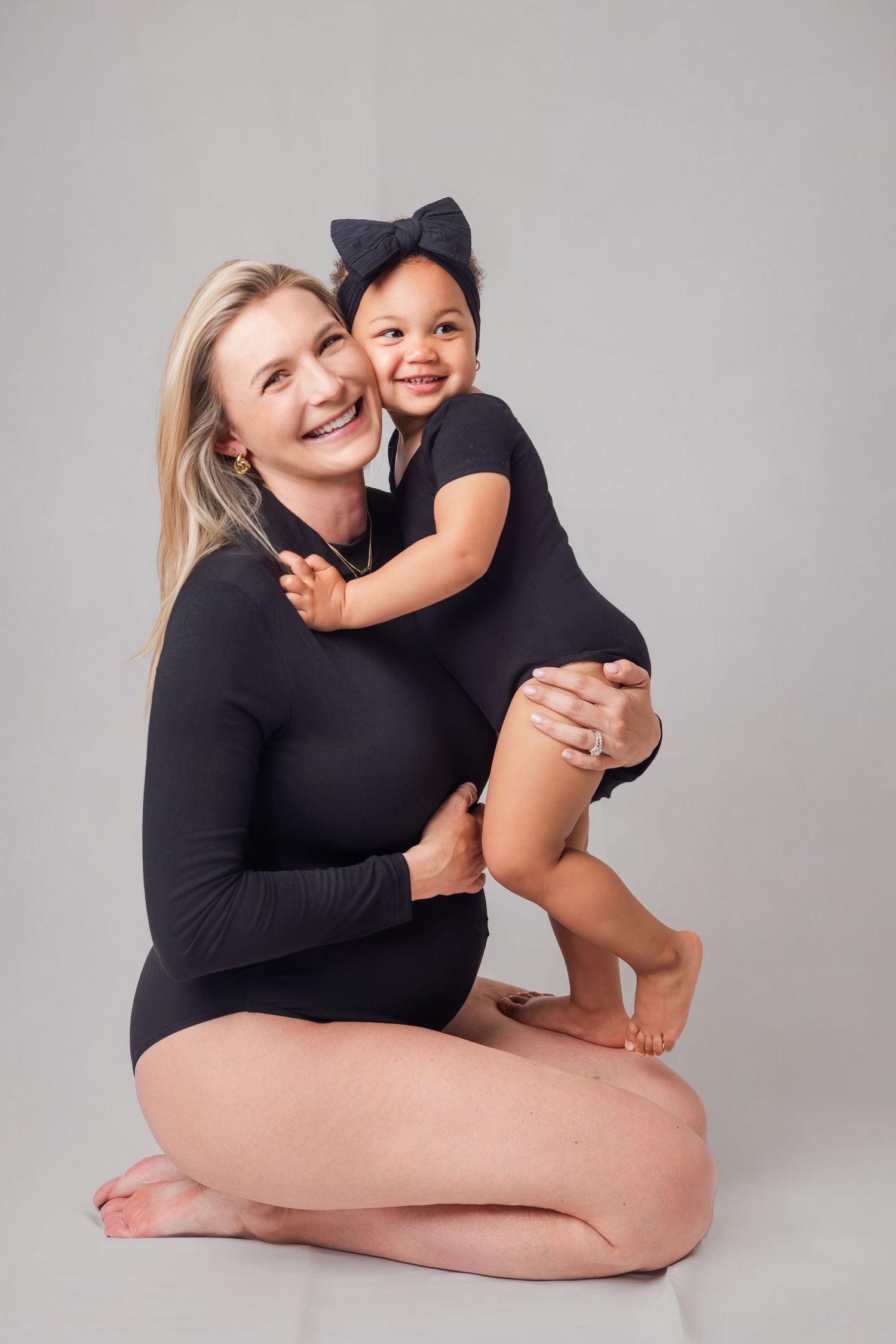 Pregnant blonde woman in black bodysuit hugs smiling toddler daughter in studio.