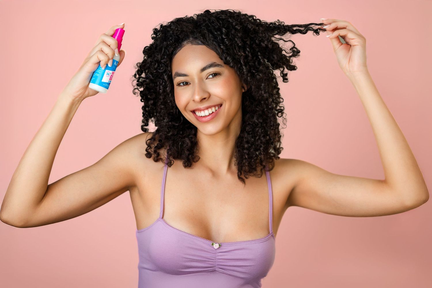 Woman with curly hair holding blue spray bottle styling product in purple bralette.