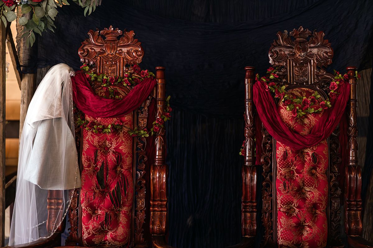 Bride in white veil kneeling before ornate red ceremonial chairs decorated with flowers and red fabric.