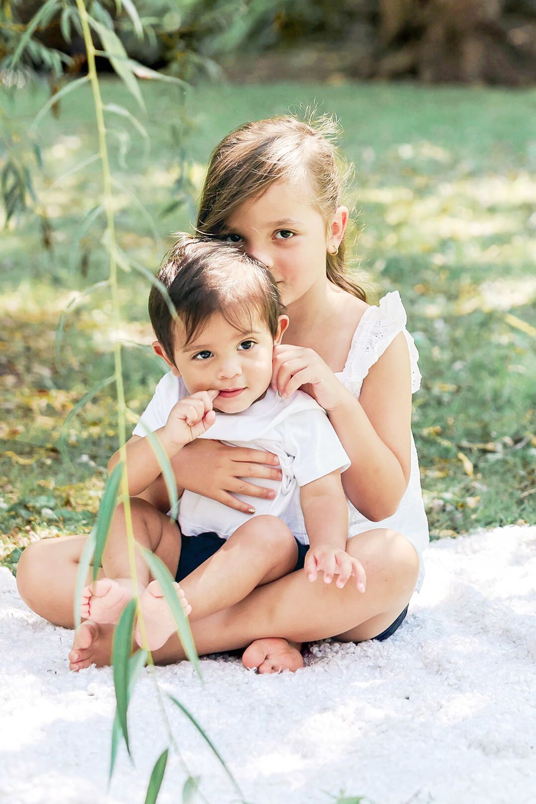 A young girl lovingly hugs her baby sibling while sitting on a blanket in a lush green park.