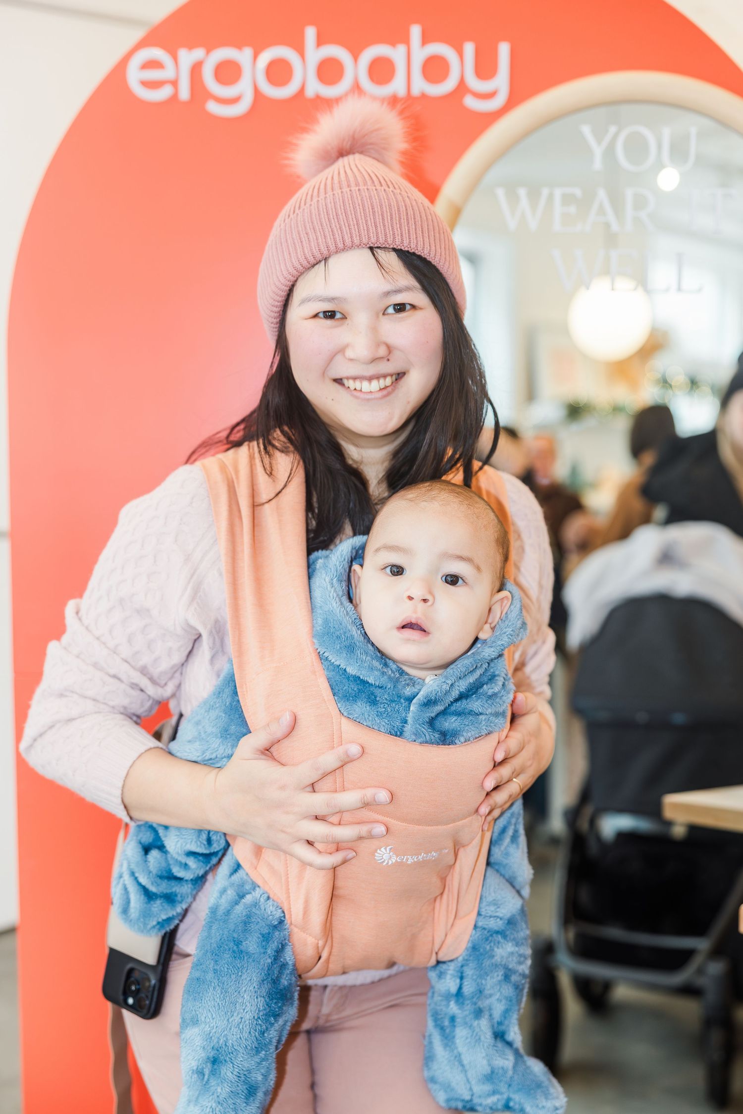 Smiling mother wearing pink beanie holds baby in Ergobaby carrier at branded event.