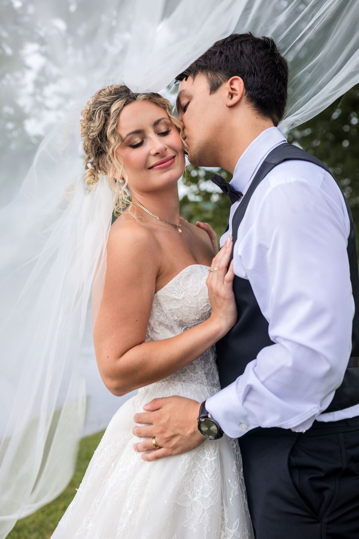 Bride and groom share a romantic moment, groom kissing bride on cheek, with flowing white fabric backdrop outdoors.