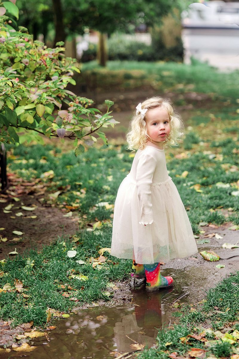 A blonde toddler girl in a white tulle dress and rainbow rain boots plays in a fall park puddle.