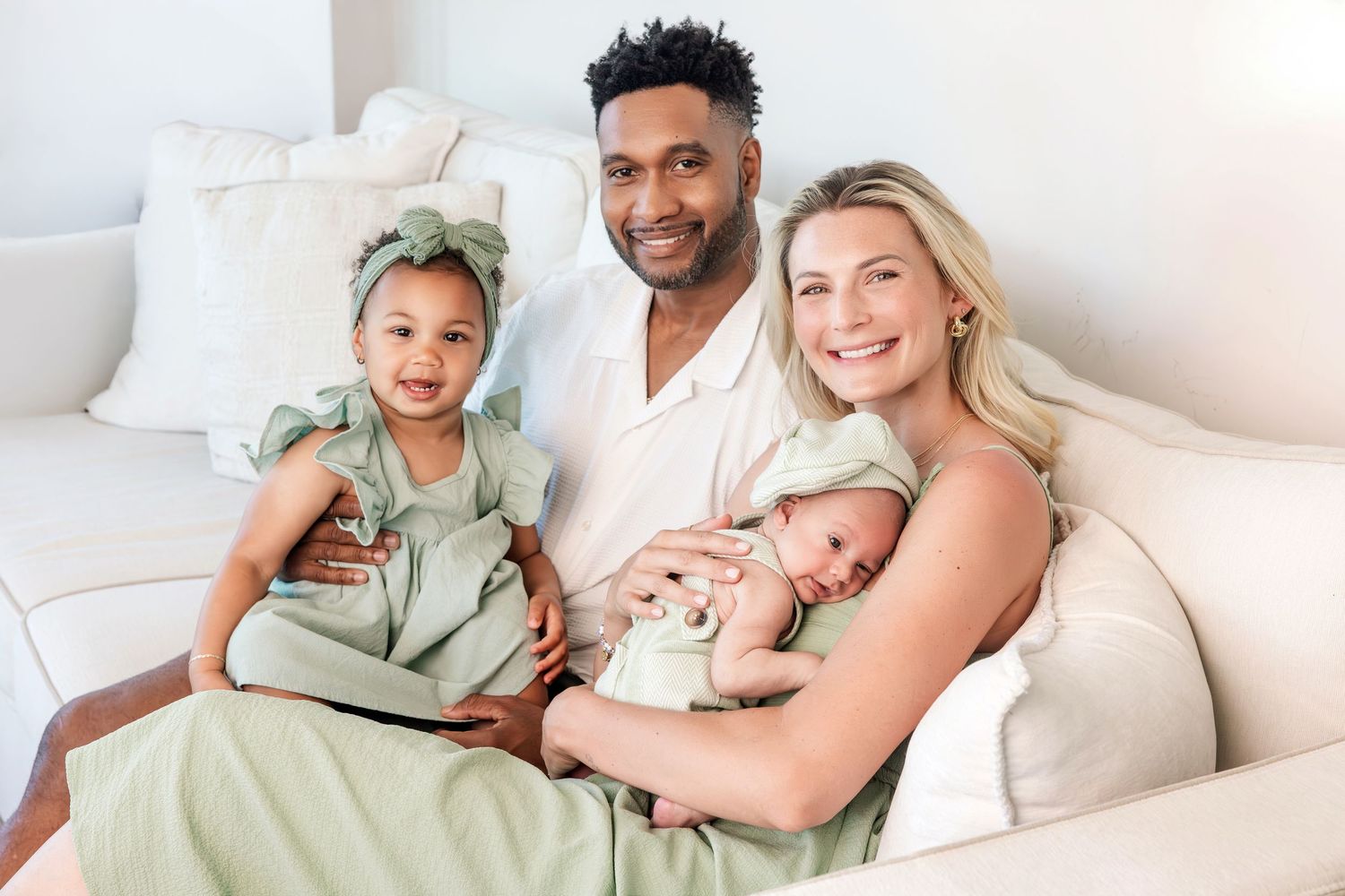 Happy interracial family of four with toddler and newborn sitting together on white sofa.