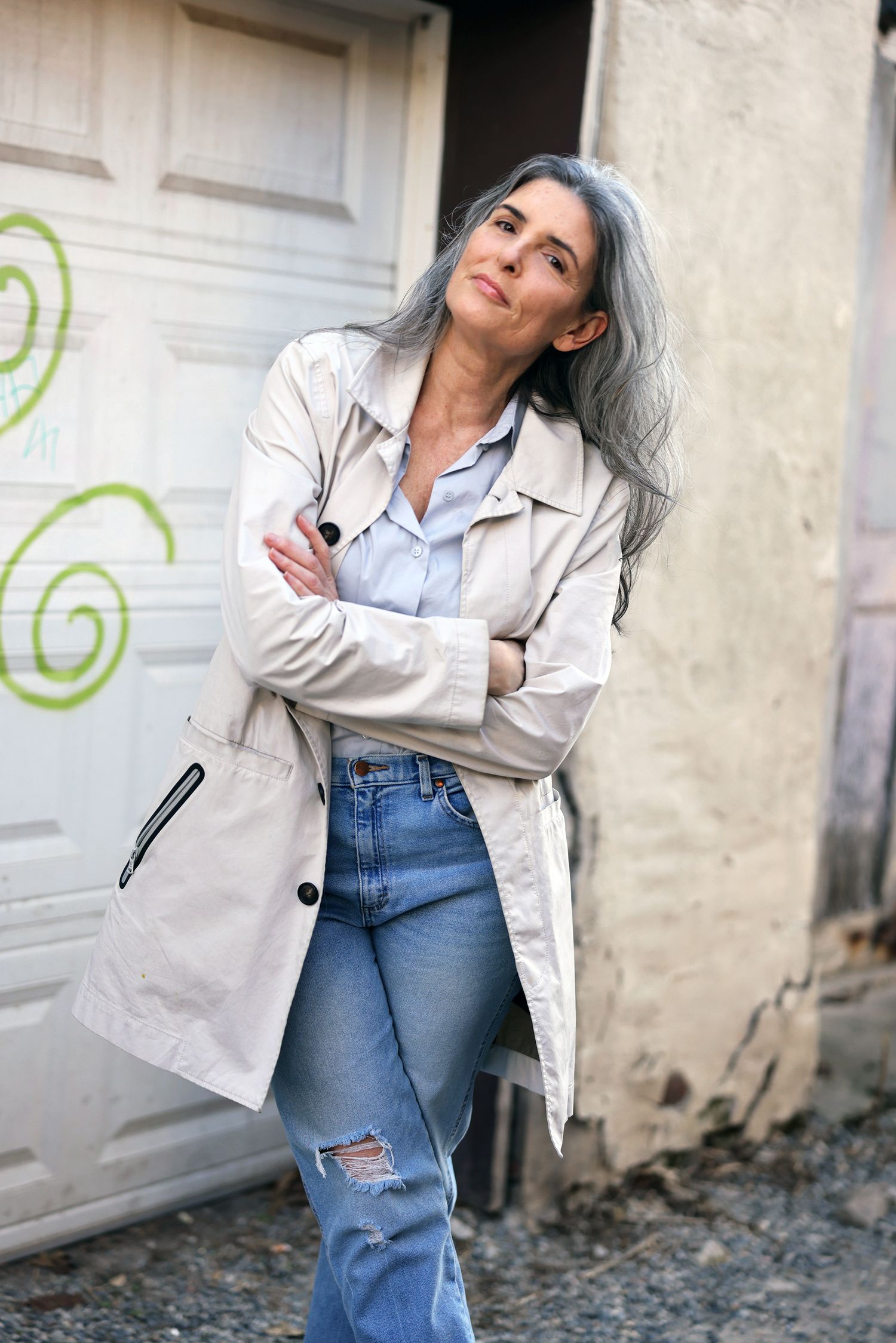 A stylish woman with long gray hair poses in ripped jeans and a beige trench coat, arms crossed, leaning against a wall.
