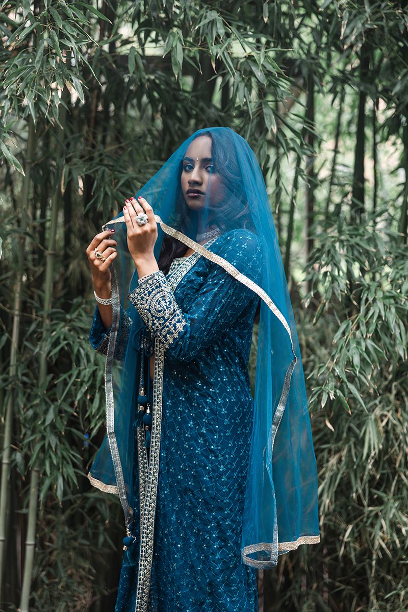 Woman in teal embroidered salwar kameez with matching dupatta veil poses gracefully among bamboo trees.