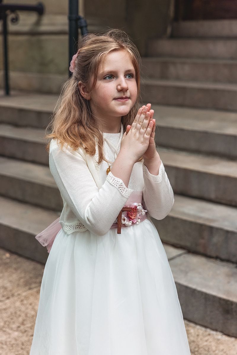 Young girl in white First Communion dress with hands clasped in prayer on church steps.