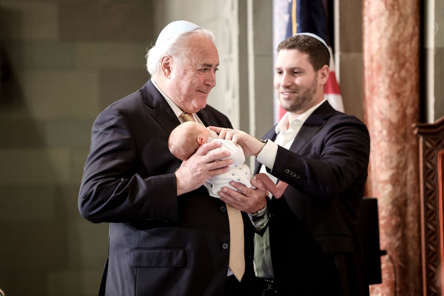Older man in suit holds newborn baby while younger man smiles during Jewish ceremony.