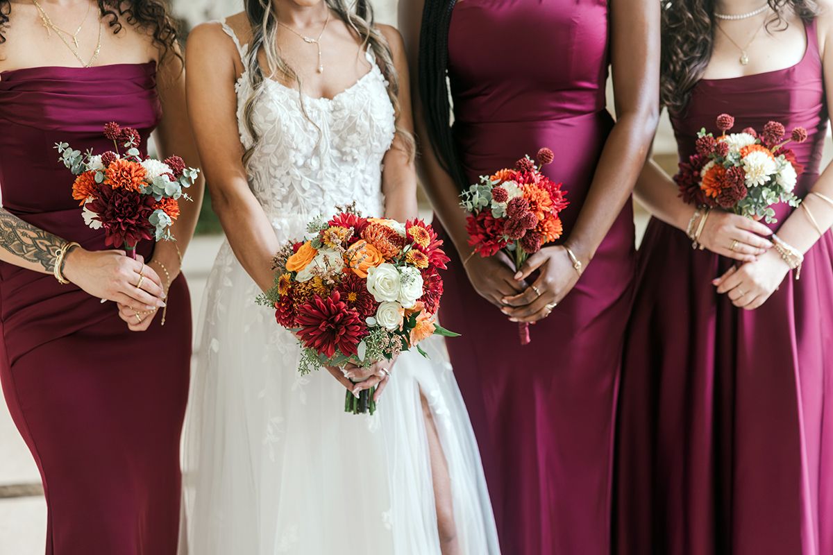 Bride in white gown with bridesmaids in burgundy dresses holding red and orange bouquets at wedding ceremony.