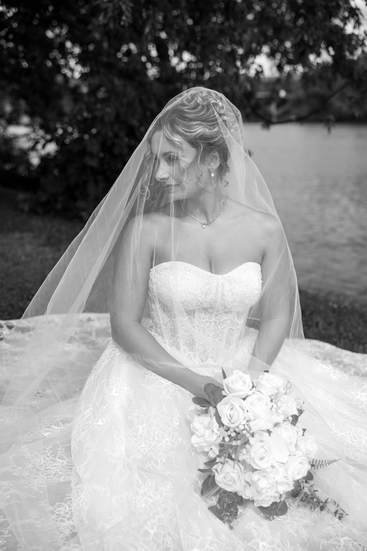 Black and white bridal portrait of a bride in a strapless lace gown, veil, holding a rose bouquet outdoors.