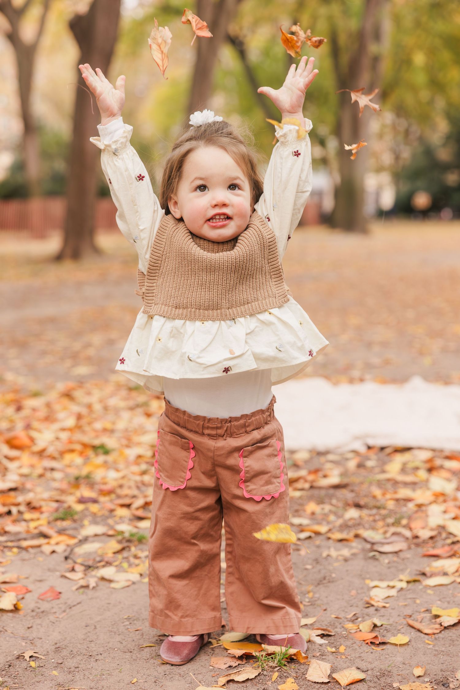 Toddler girl in autumn park tossing fall leaves in the air, wearing brown corduroy pants and knit vest.