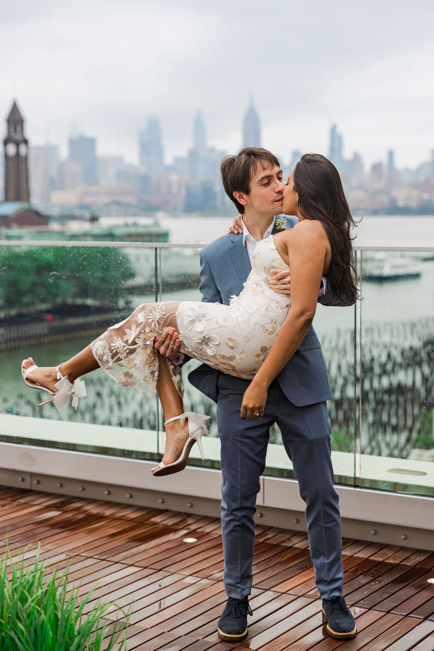 Groom lifting bride in floral dress for a kiss on rooftop deck with NYC skyline behind them.