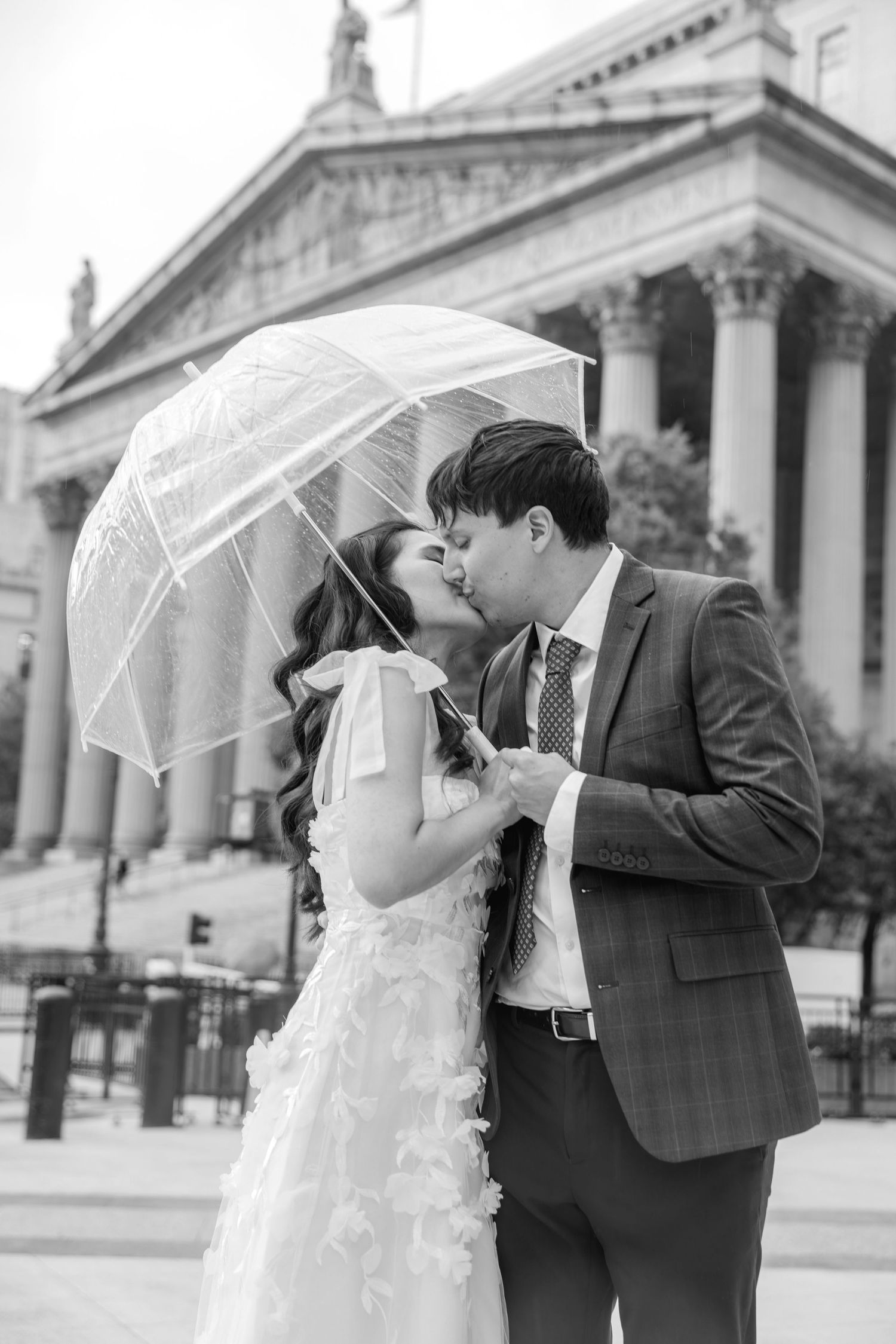 Black and white photo of a bride and groom kissing under a clear umbrella outside a grand building.