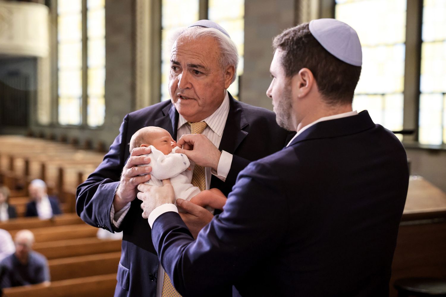 Two men in suits and kippahs carefully transfer newborn baby during Jewish bris ceremony.