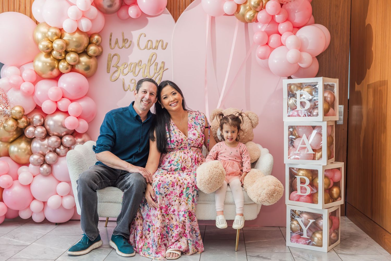 Family at a pink baby shower with balloon arch, pregnant mother, father, and toddler daughter on swing.