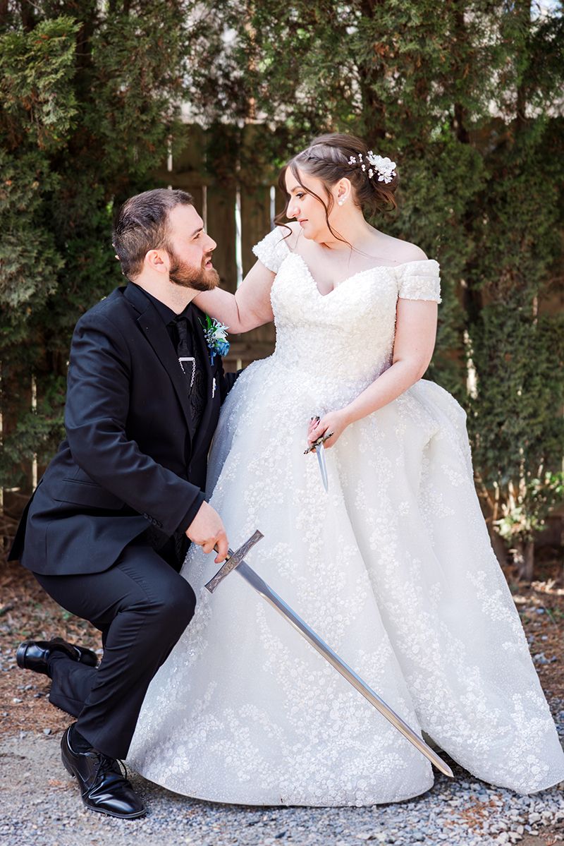 Bride in lace ball gown and groom in black suit pose with a sword in a romantic fantasy-themed wedding.
