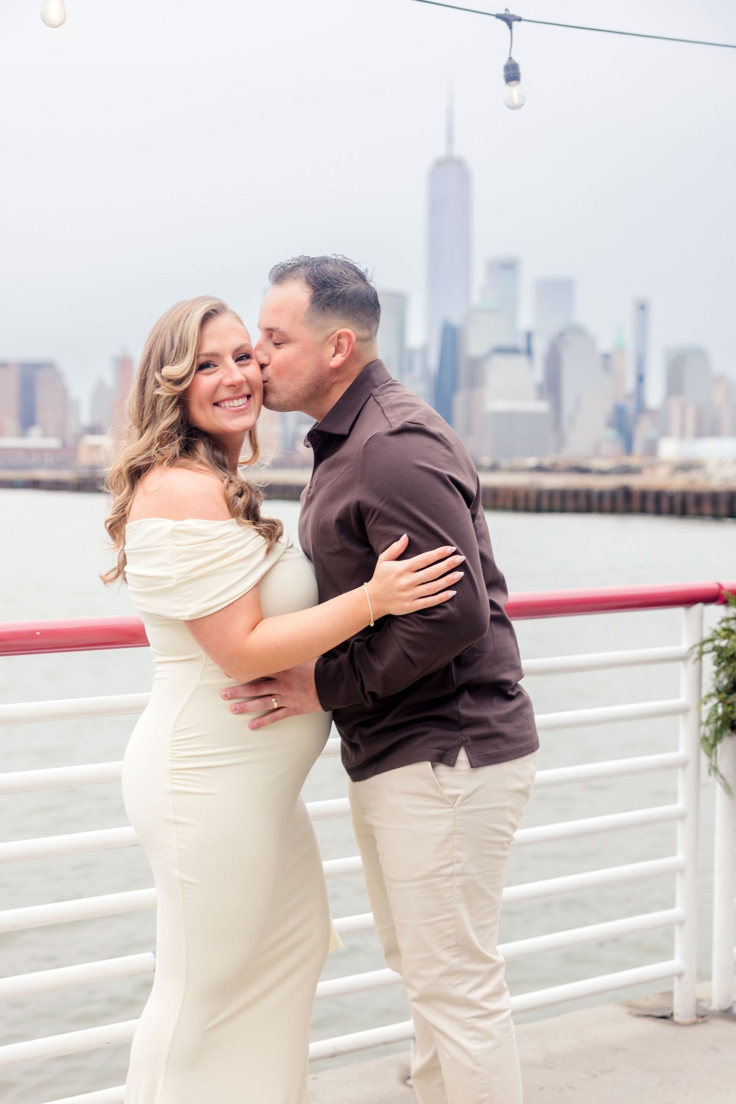 Pregnant woman in white off-shoulder gown receives kiss from partner with NYC skyline backdrop.