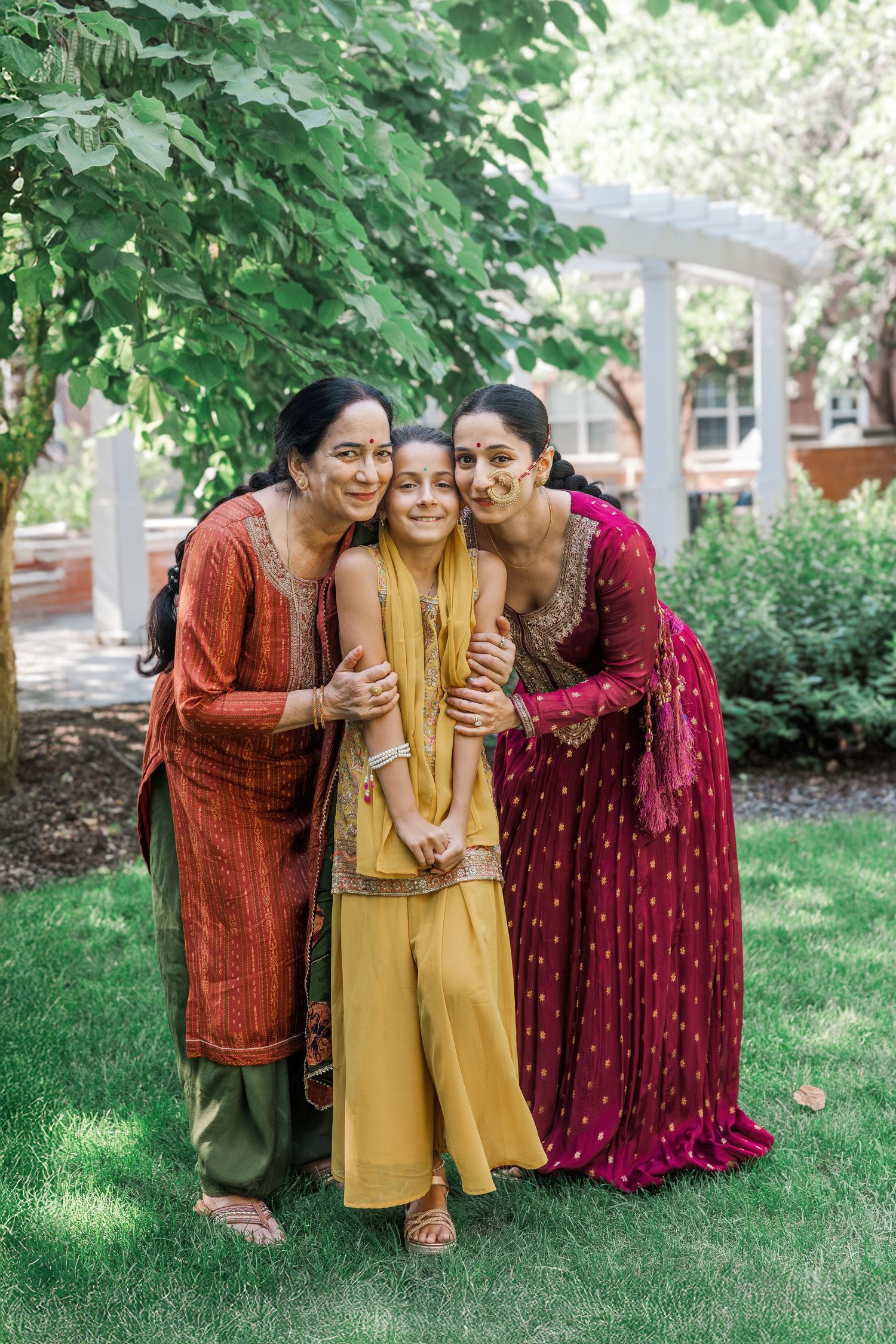 Three women in colorful traditional Indian attire smile and embrace outdoors in a lush garden setting.