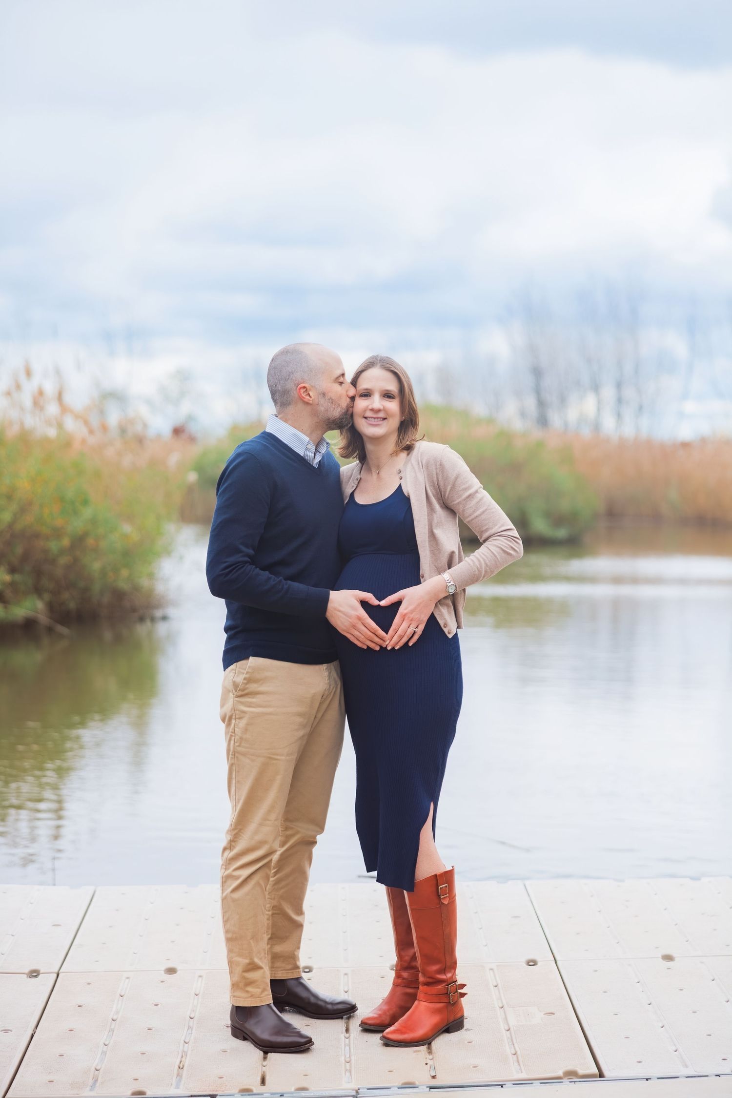 Couple standing on dock by lake, man kissing pregnant woman's cheek as they form heart shape over baby bump.
