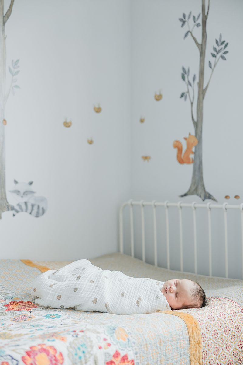 Newborn baby swaddled in white muslin blanket sleeping peacefully in a crib with woodland nursery decor.