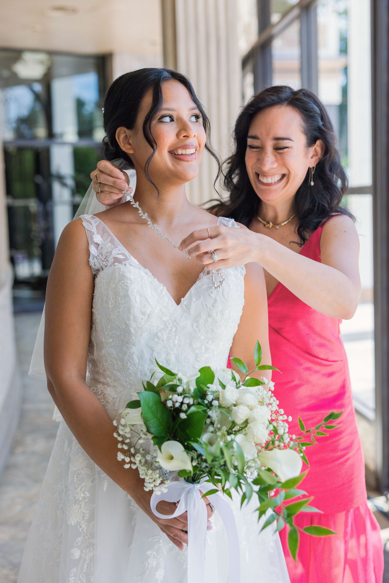 Bride in lace wedding dress holding white bouquet while woman in pink dress adjusts her veil outdoors.