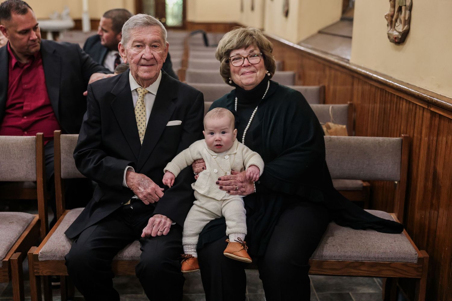 Elderly grandparents sit on church pew smiling with baby grandchild on lap.