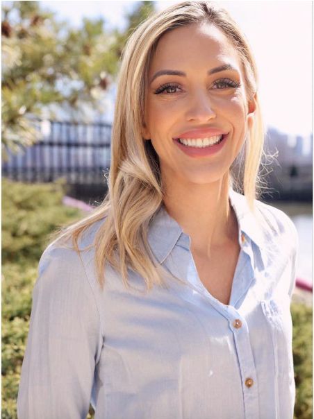 Smiling blonde woman in a light blue button-up shirt posing outdoors with greenery and a city skyline.