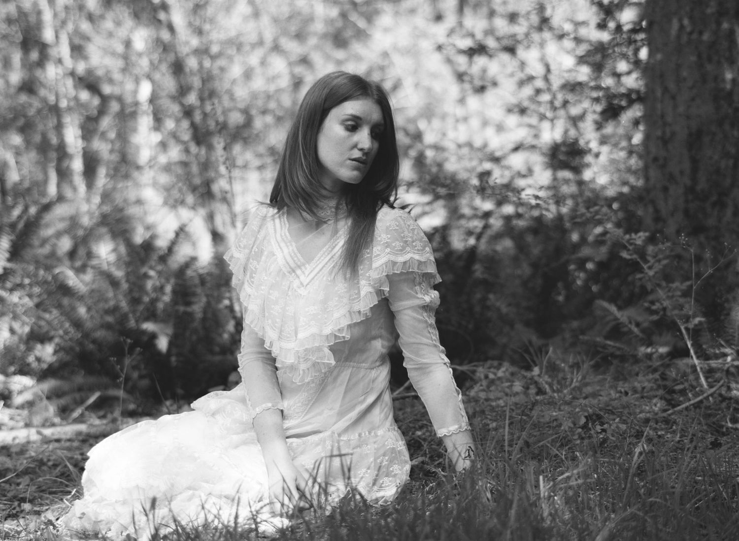 Black and white portrait of young woman in white lace dress sitting in forest setting.