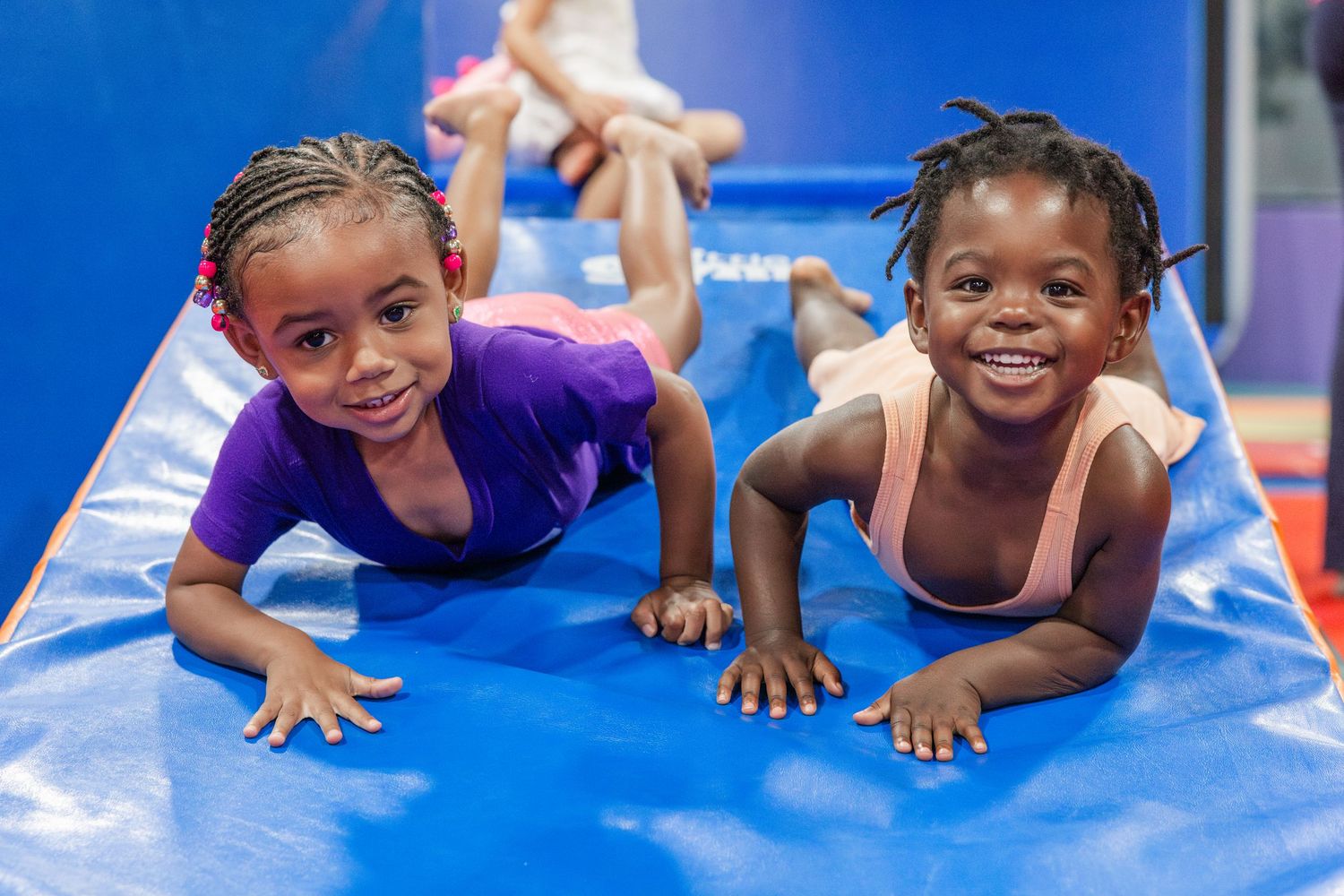 Two young girls in leotards smile while doing planks on a blue gymnastics mat in a colorful gym.