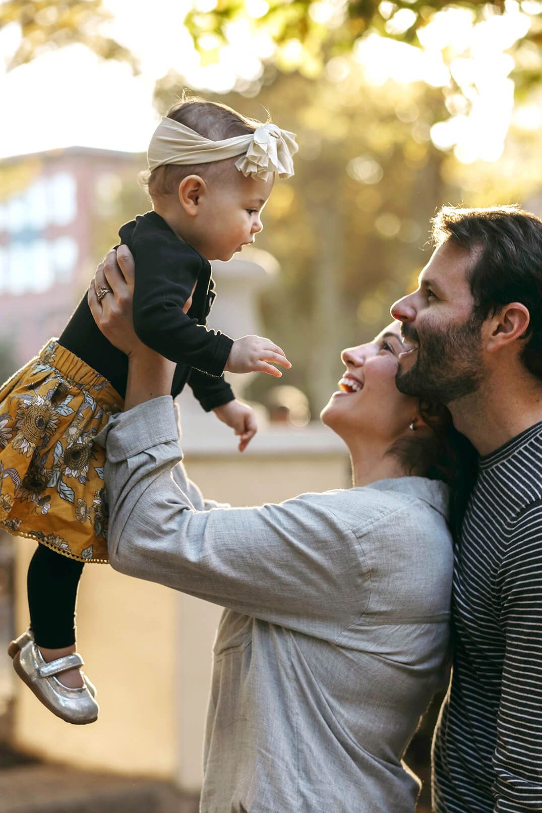 Happy parents lift their adorable baby girl outdoors in warm golden sunlight among autumn trees.