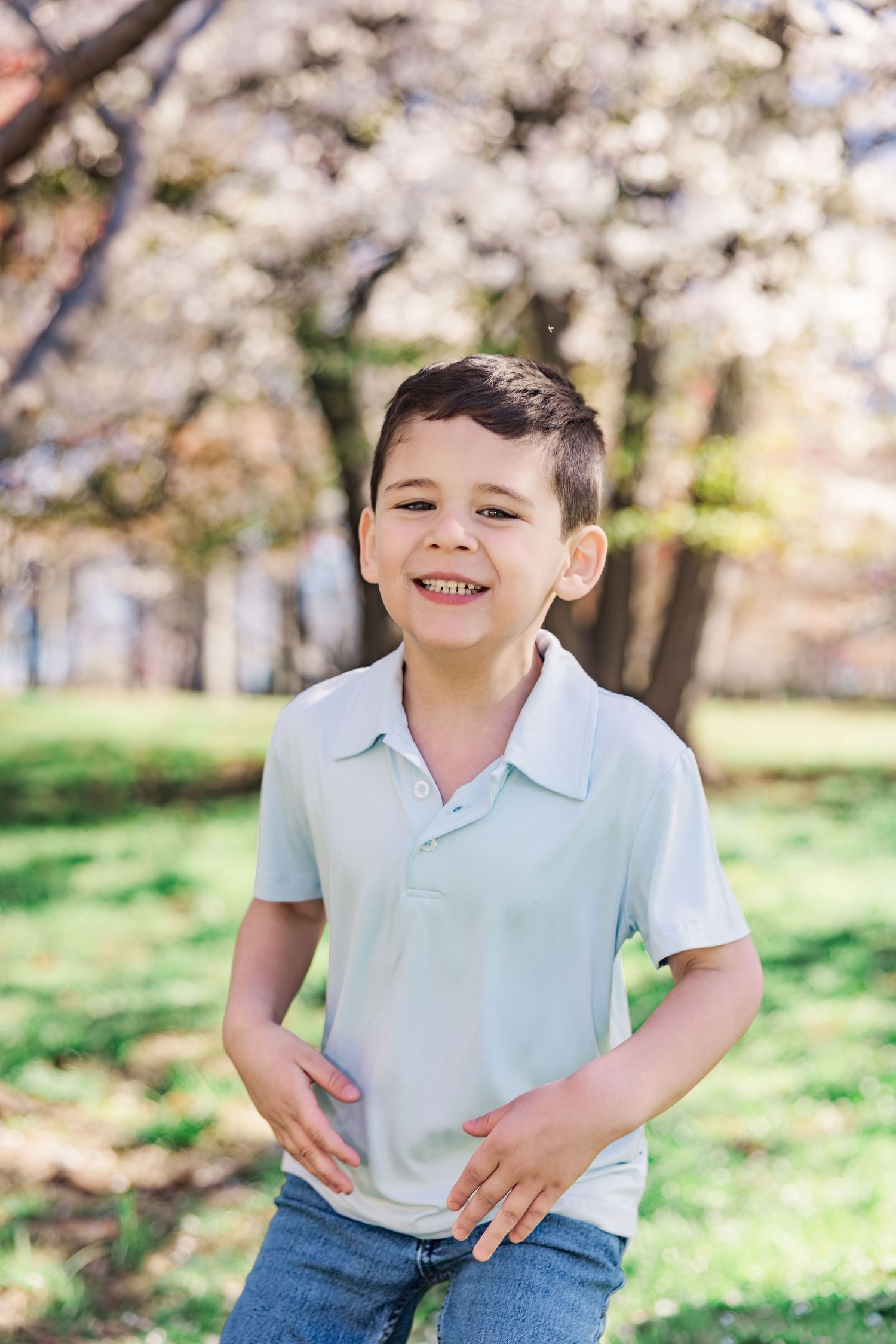 A happy young boy in a light blue polo shirt smiles in a sunny park with blossoming trees.