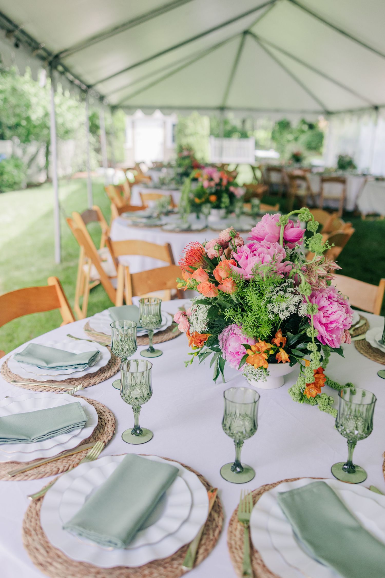 Elegant outdoor wedding reception table with green goblets, woven chargers, sage napkins, and vibrant pink and orange floral centerpiece.