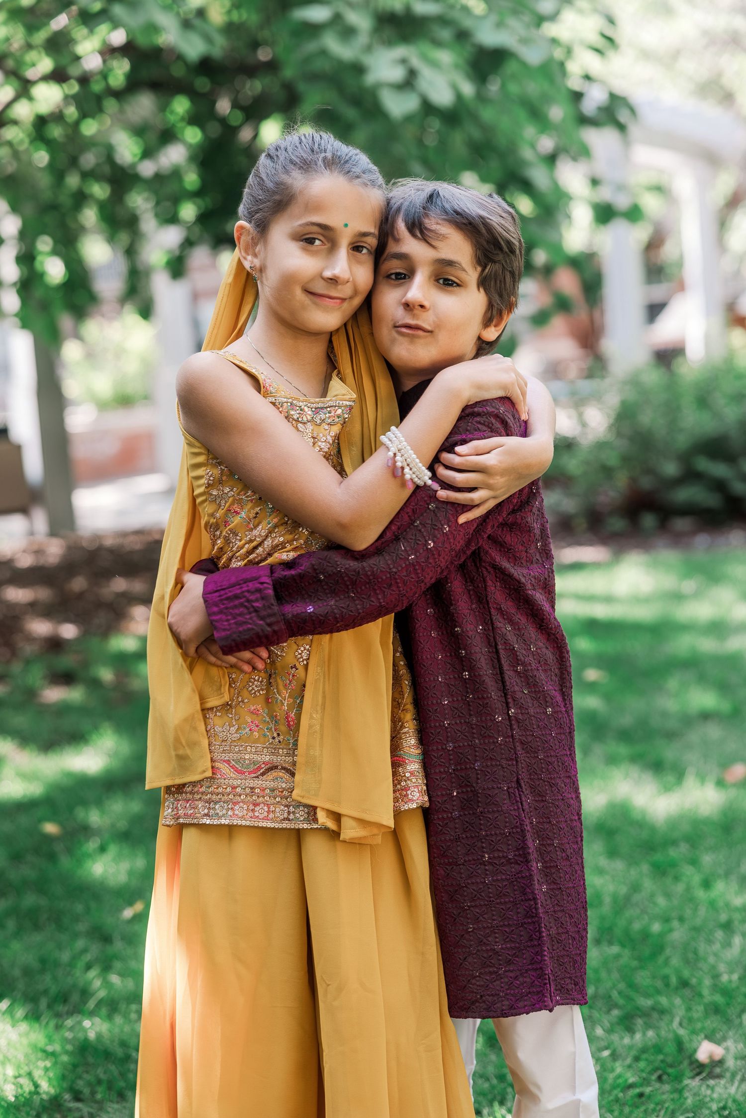 Two children in traditional South Asian attire embrace warmly outdoors, wearing gold and maroon embroidered outfits.