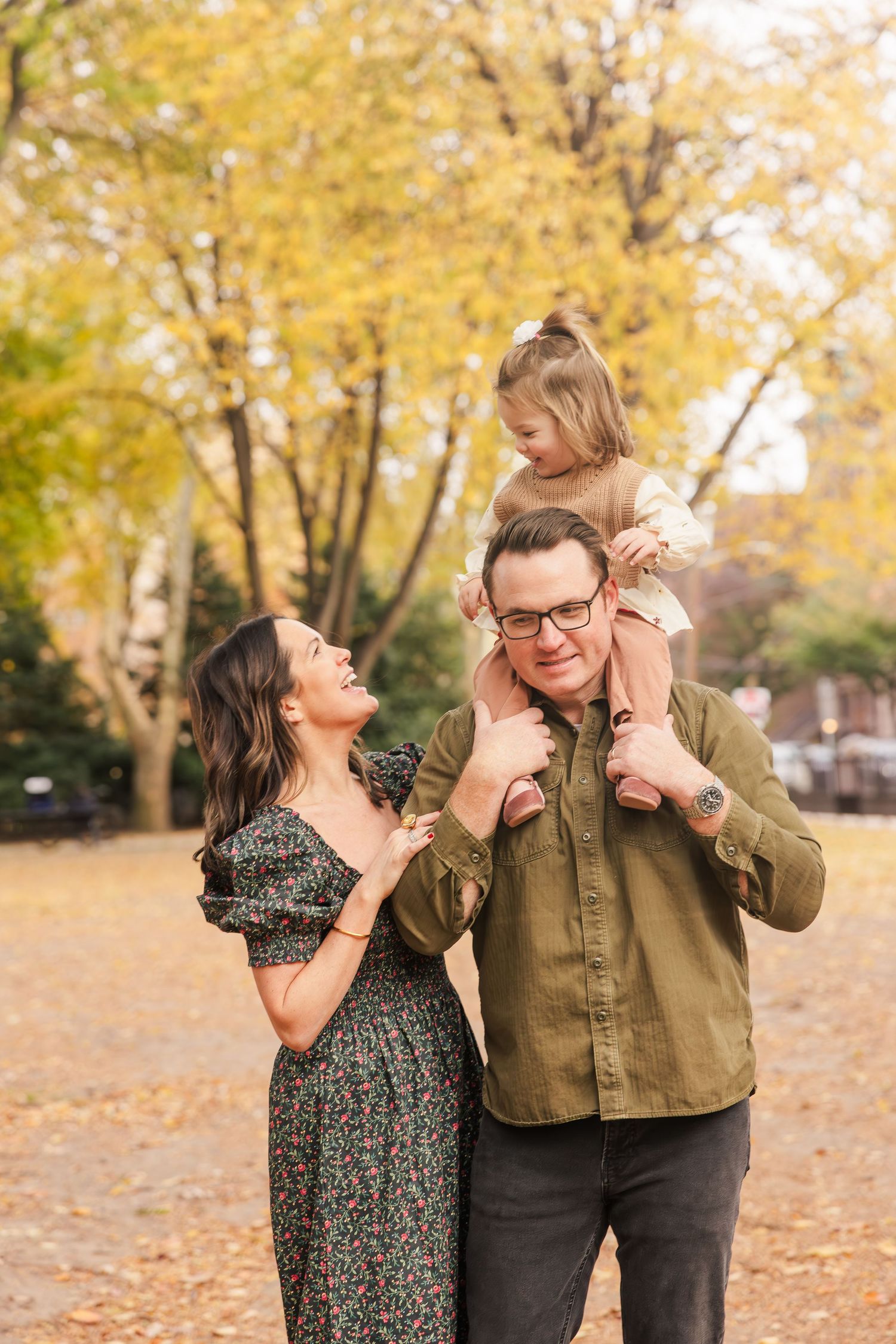 Happy family of three in autumn park, dad carrying toddler on shoulders while mom laughs beside them.