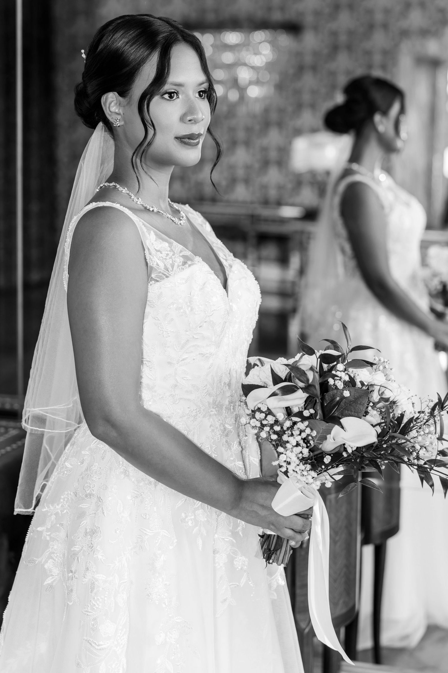 Elegant bride in lace wedding gown holding floral bouquet with veil, black and white portrait photo.
