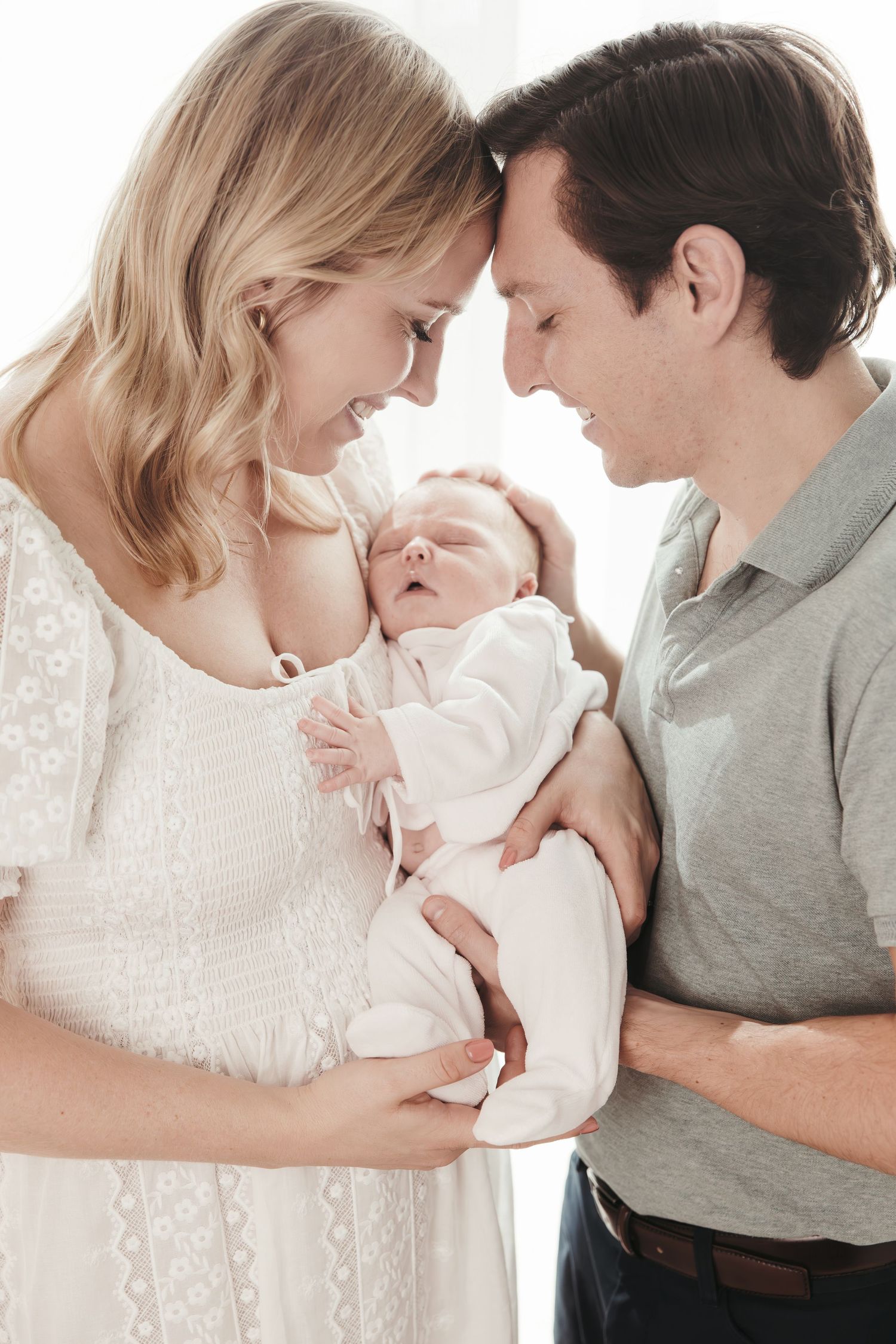 New parents touching foreheads and smiling while holding sleeping newborn baby near bright window.