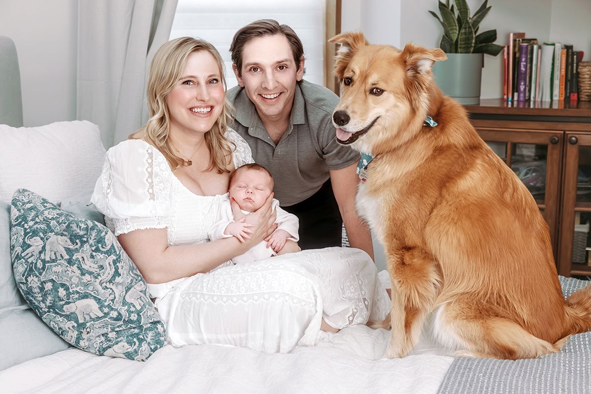 Happy family portrait with mother, father, newborn baby, and large fluffy brown dog on white sofa.