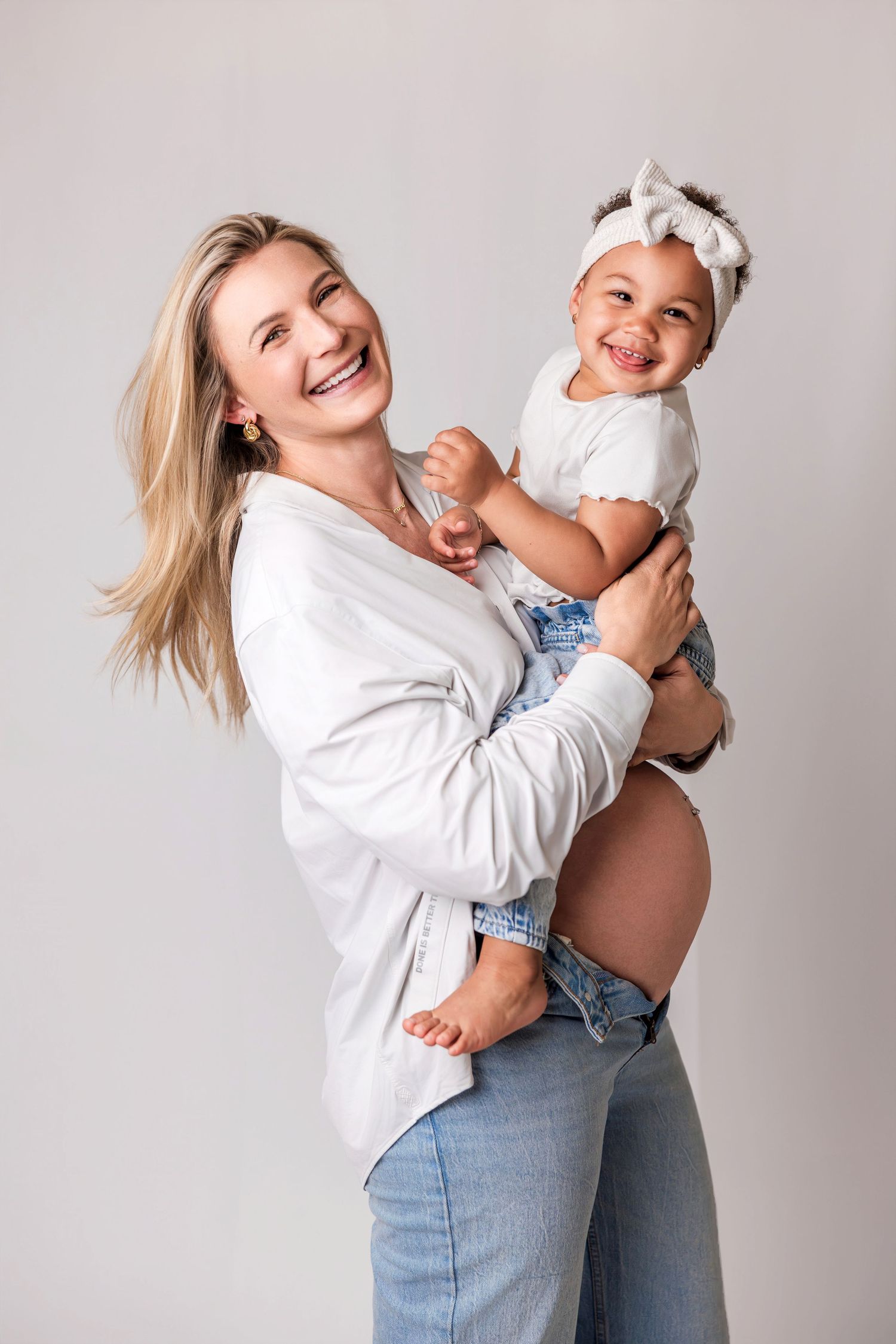 A joyful pregnant woman in a white shirt holds a laughing toddler girl with a white bow headband.