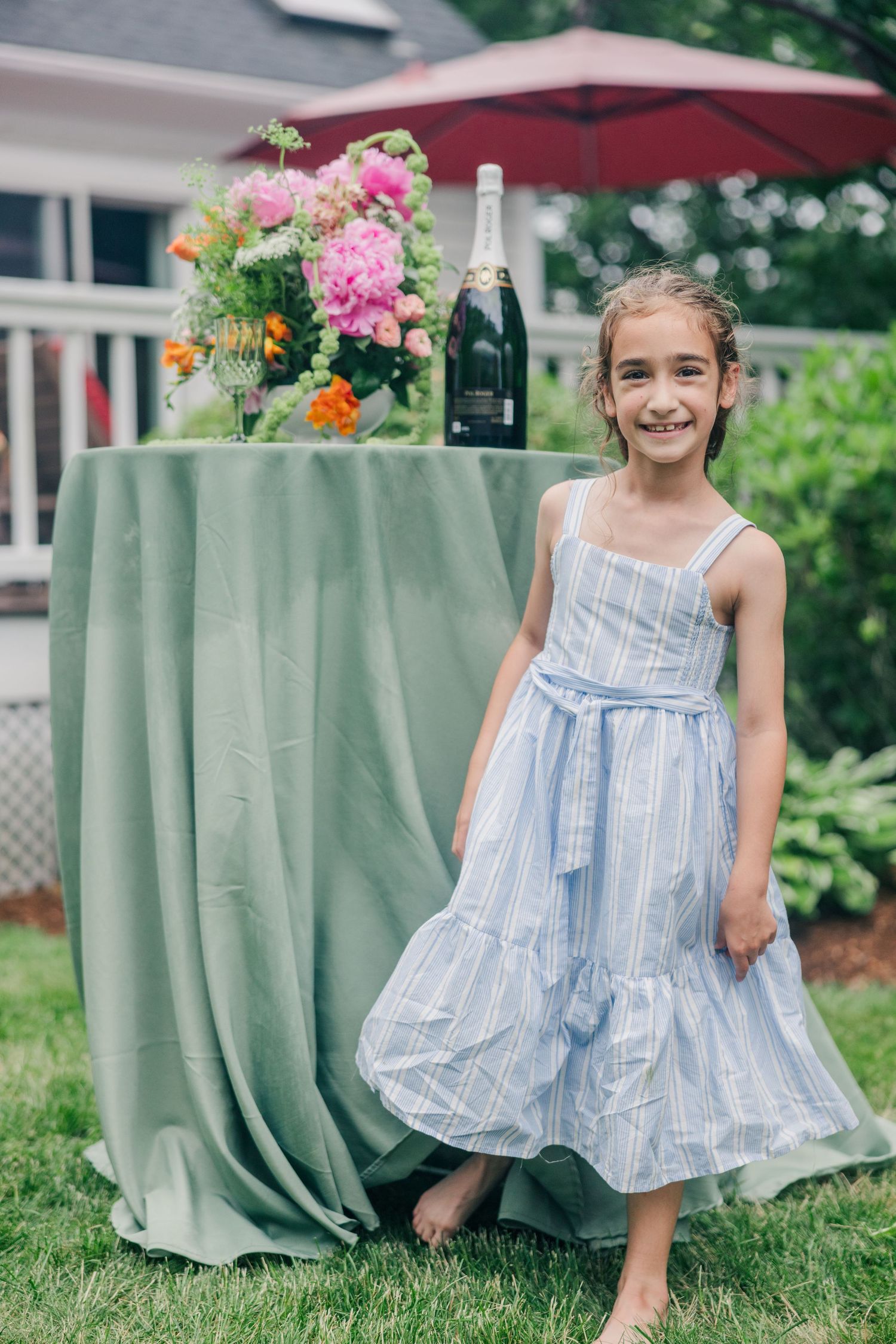 Young girl in blue striped dress smiling outdoors near floral arrangement and champagne bottle on green draped table.