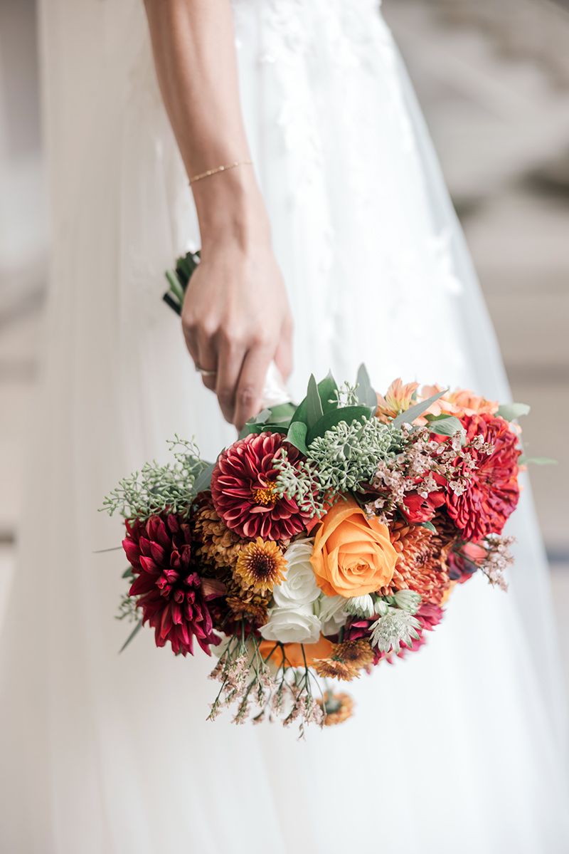 Bride in white gown holding a vibrant fall wedding bouquet with red dahlias, orange roses, and white blooms.