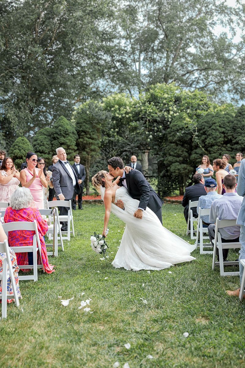 Bride and groom share a romantic dip kiss in the aisle during an outdoor garden wedding ceremony.