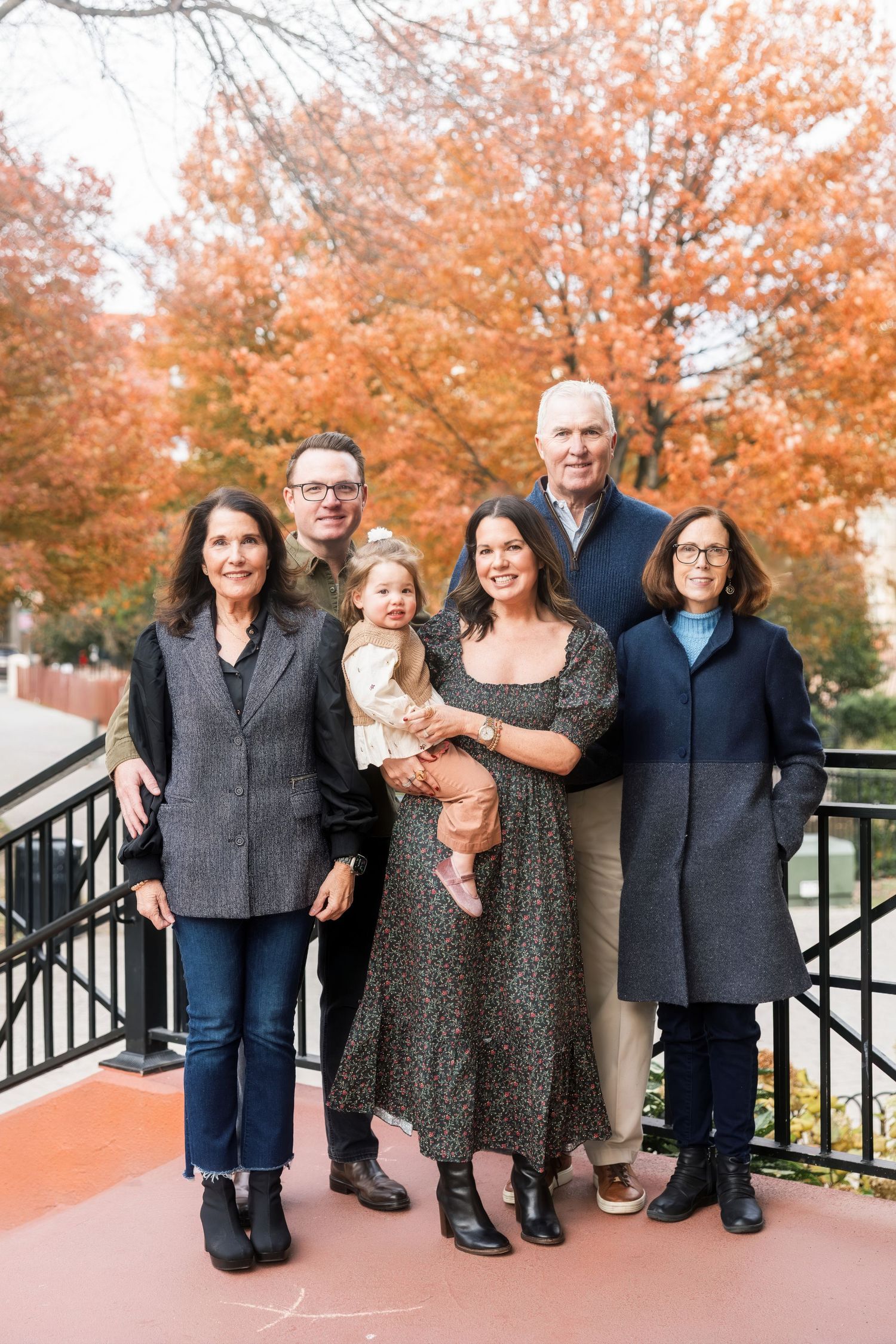 Multi-generational family of six posing outdoors in front of vibrant red autumn foliage.