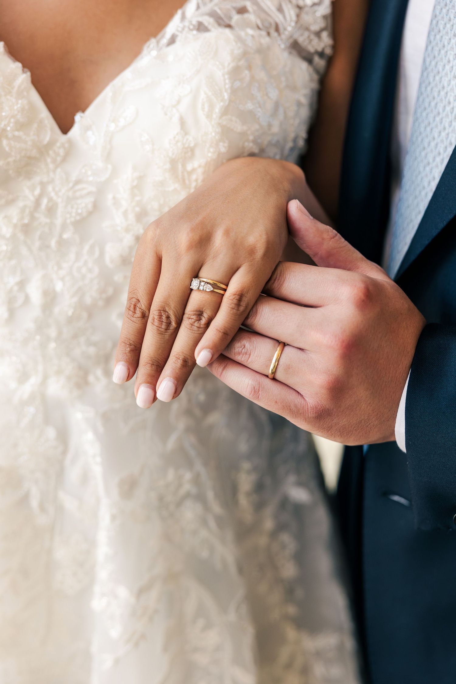 Close-up of newlywed couple showing gold wedding rings, bride in lace dress, groom in navy suit.
