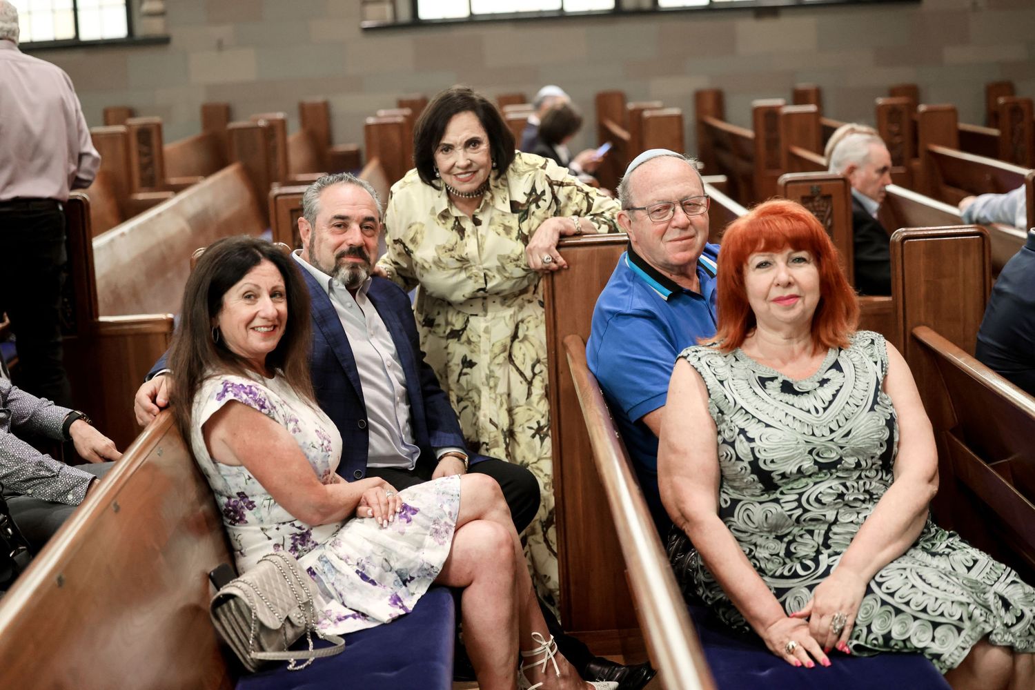 Group of five adults smiling together while seated in synagogue pews at Jewish ceremony.