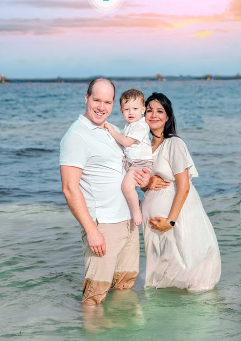 Happy family with toddler stands in ocean water at sunset during maternity photo session.