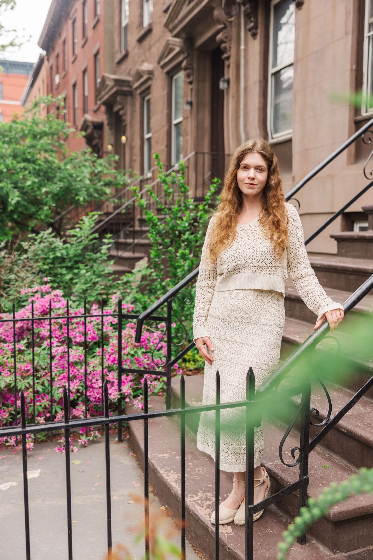 Elegant woman in white knit dress poses on historic brownstone steps surrounded by pink blooming flowers.